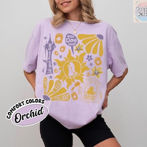 K&ouml;nnte beinhalten: Ein helllila T-Shirt mit einem gelben Grafikdesign mit einer Sonne, Blumen und Text. Der Text enth&auml;lt "best day ever", "Let your Power Shine" und "At Last I See the Light". Das Shirt ist mit "Comfort Colors Orchid" gekennzeichnet.