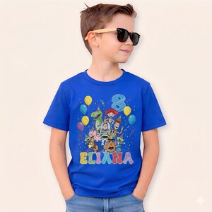 May include: A royal blue t-shirt featuring a colorful graphic of Toy Story characters, balloons, confetti, the number 8, and the name "ELIANA" in rainbow letters. The child is wearing sunglasses.