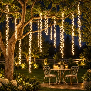 Op de afbeelding: Meerdere strengen warmwitte LED-lichtjes hangen verticaal aan de takken van een grote boom. Onder de boom staat een ronde metalen tafel met twee bijpassende stoelen op een terras, verlicht door kleine kaarsjes in glazen houders.