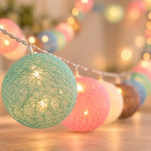May include: A string of decorative fairy lights featuring spherical shades made of interwoven thread in various colours including mint green, bright pink, white, and brown, with warm internal illumination glowing through the textured spheres.