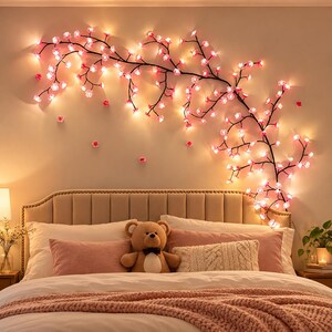 Peut inclure: Une décoration lumineuse murale en forme de branche de cerisier en fleurs, dotée de tiges sombres et de nombreuses petites lumières LED roses en forme de fleurs. La branche s'étend horizontalement sur le mur au-dessus d'un lit.