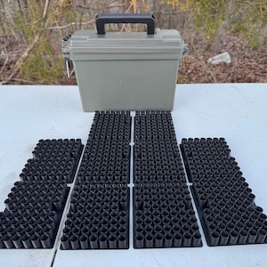 May include: An olive green storage container with a black handle sits above several black foam inserts. The inserts are designed to hold small cylindrical objects, likely ammunition or similar items. The scene is set outdoors.