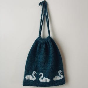 Op de afbeelding: Een tealgebreide trekkoordtas met drie witte zwanen geborduurd langs de onderkant. De tas hangt tegen een witte muur. Het trekkoord van de tas is gemaakt van hetzelfde teal garen.