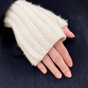 May include: Cream-colored fingerless gloves with a ribbed knit design. The gloves are worn over a dark navy blue coat. The gloves are made of a soft, warm material, perfect for cold weather.