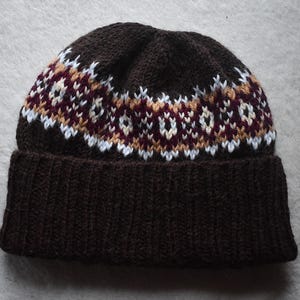 May include: A dark brown knit beanie with a patterned design in white, burgundy, and tan. The hat features a ribbed brim and a fair isle pattern across the crown. This winter hat is perfect for cold weather.