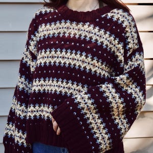 Pode incluir: Um suéter bordô com um padrão geométrico em creme, azul claro e marrom. O suéter tem gola redonda e mangas compridas. Os punhos e a bainha são canelados.