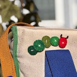 May include: Beige canvas bag with a blue strap and an orange shoulder strap. The bag is decorated with felt circles in green and red, resembling a caterpillar. The green circles have white cross stitches.