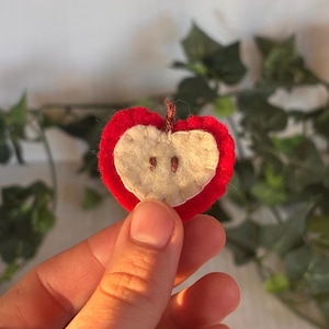 May include: A small, heart-shaped felt apple ornament featuring a bright red outer layer and a cream-colored center. The center piece is stitched with two small brown vertical lines representing seeds and includes a tiny brown stem at the top.