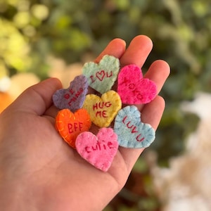 May include: A hand holds a collection of colorful felt heart-shaped ornaments. Each heart is a different color, including pink, orange, yellow, and blue, with embroidered phrases like "BFF", "LUVU", and "HUG ME". The hearts are a charming craft item.