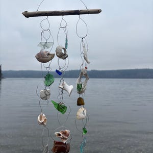 May include: A handmade wind chime with a driftwood base and wire strands suspending natural elements. These include seashells, sea glass in green, blue, and clear, and other organic shapes. The chime hangs against a backdrop of a lake and overcast sky.