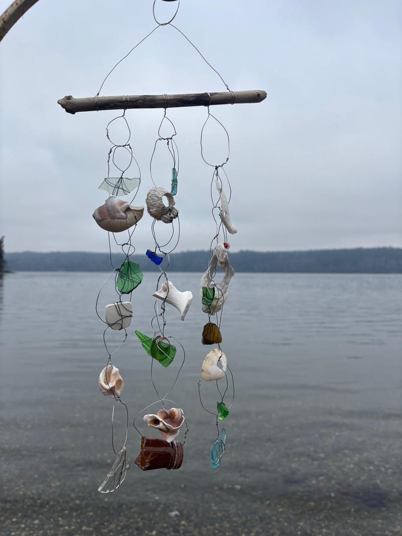 May include: A handmade wind chime with a driftwood base and wire strands suspending natural elements. These include seashells, sea glass in green, blue, and clear, and other organic shapes. The chime hangs against a backdrop of a lake and overcast sky.
