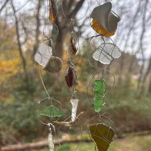 Pode incluir: Um sino de vento artesanal com várias formas de vidro do mar em tons de verde, amarelo, castanho e branco, unidos por fio de prata. As peças de vidro do mar são suspensas de uma série de laços de arame, criando uma estética decorativa e natural.