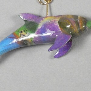 May include: A pair of handcrafted dolphin earrings. Each earring features a colorful dolphin with purple fins, a blue tail, and a gold-colored hook. The dolphins have a painted design with green, brown, and yellow accents.