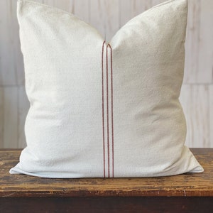 May include: A square throw pillow made of off-white, textured fabric. The pillow features a vertical design element consisting of three thin, parallel dark red stripes running down the center of the front face from top to bottom.