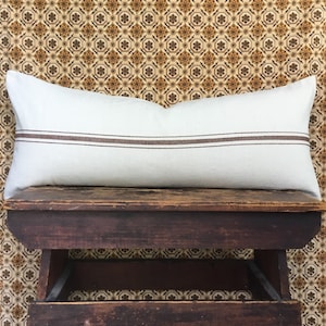 May include: A white lumbar pillow with two brown stripes on a wooden stool. The stool is sitting on a patterned floor.