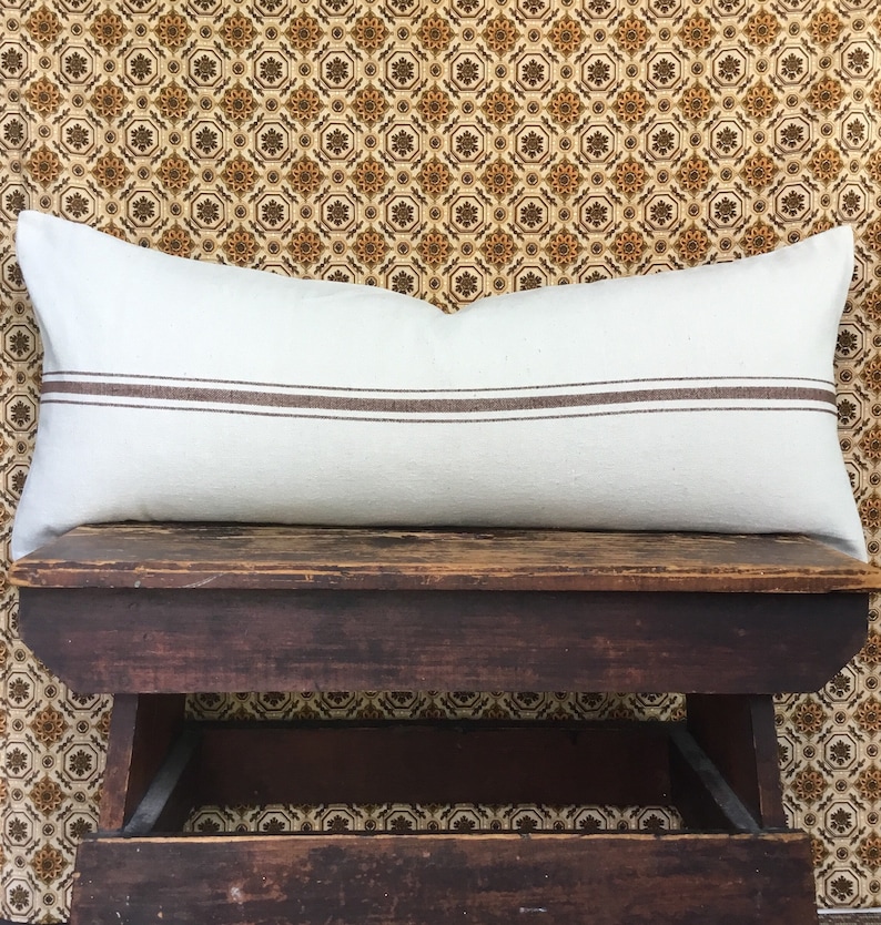 May include: A white lumbar pillow with two brown stripes on a wooden stool. The stool is sitting on a patterned floor.