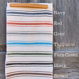 May include: A white fabric with colorful stripes in navy, red, gray, turquoise, fern green, black, brown, mustard, and ochre. The fabric is hanging on a wooden hanger.