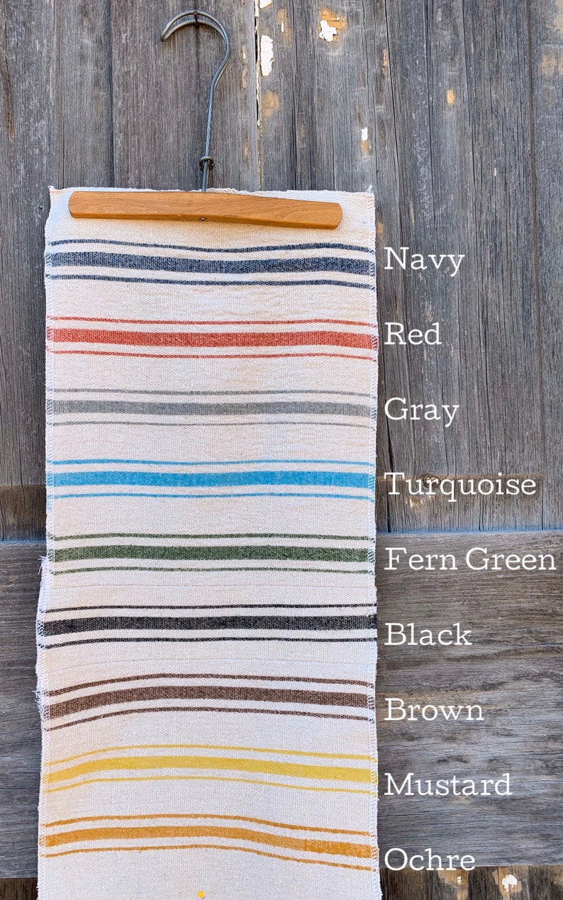 May include: A white fabric with colorful stripes in navy, red, gray, turquoise, fern green, black, brown, mustard, and ochre. The fabric is hanging on a wooden hanger.