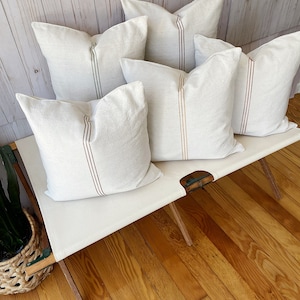 May include: Five square throw pillows made of off-white fabric, each featuring a single vertical stripe running down the center. The stripes vary in color, including shades of dark green, brown, rust, and mustard yellow, set against the light fabric.