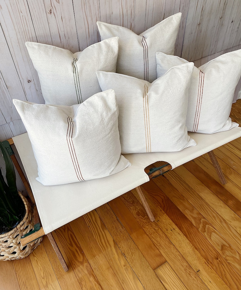 May include: Five square throw pillows made of off-white fabric, each featuring a single vertical stripe running down the center. The stripes vary in color, including shades of dark green, brown, rust, and mustard yellow, set against the light fabric.
