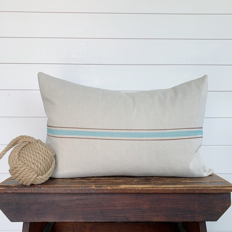 May include: A light beige throw pillow with a brown and light blue stripe design. The pillow is sitting on a wooden bench.