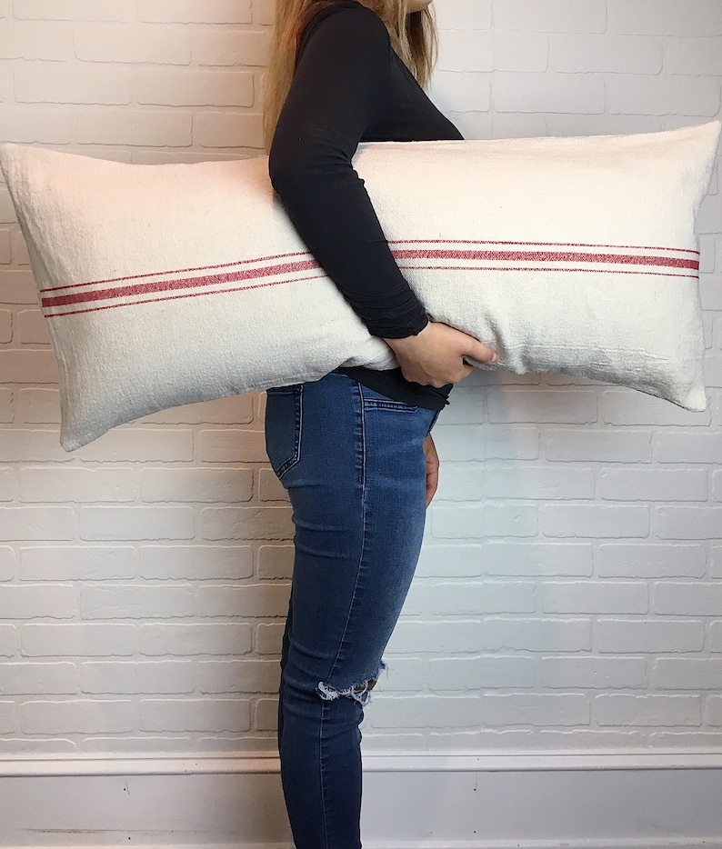 May include: A white pillow with two red stripes. The pillow is being held by a person wearing a black shirt and blue jeans.