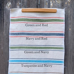May include: A white linen tea towel with five different colored stripes. The stripes are labeled with the colors: Green and Red, Navy and Red, Green and Navy, Turquoise and Navy, and Ocean Blue and Brown.