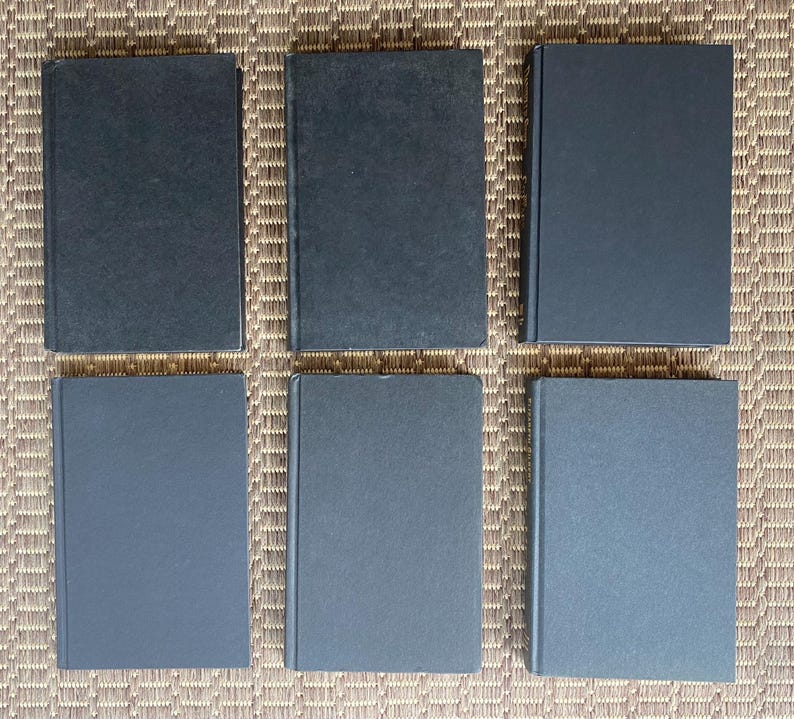 May include: Six hardcover books with dark blue covers are arranged in two rows on a textured, light brown surface. The books are rectangular and appear to be of similar size and thickness, suggesting a collection or set.