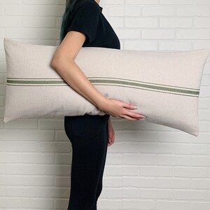May include: A person holding a long, rectangular pillow with a white linen fabric and two green stripes.