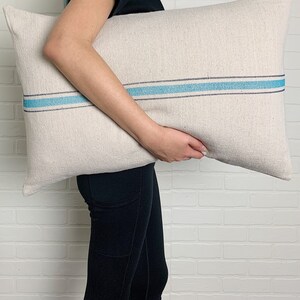 May include: A white linen pillow with a blue stripe down the center. The pillow is being held by a person wearing black pants.