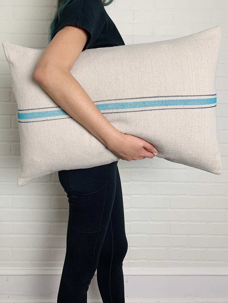 May include: A white linen pillow with a blue stripe down the center. The pillow is being held by a person wearing black pants.