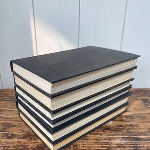 May include: A stack of six hardcover books with black covers and cream-colored pages. The books are arranged on a wooden surface, with the spines facing up. The books are in good condition.