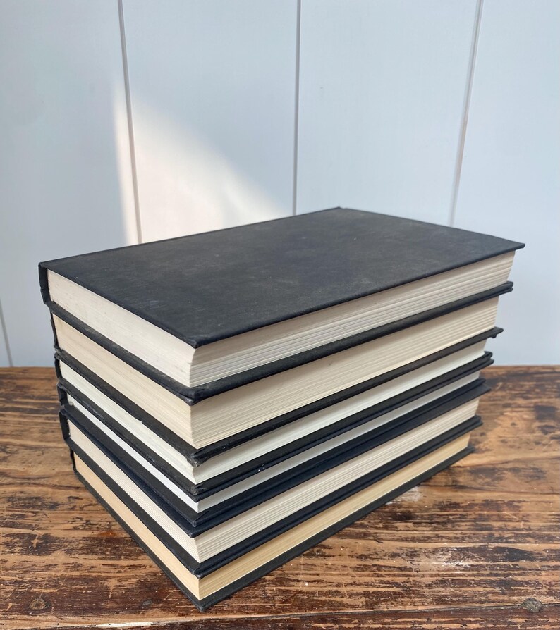 May include: A stack of six hardcover books with black covers and cream-colored pages. The books are arranged on a wooden surface, with the spines facing up. The books are in good condition.