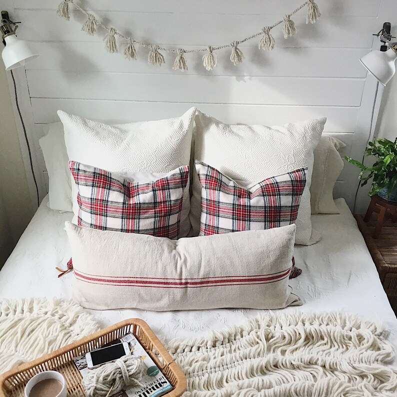 May include: A white bed with a white headboard and a white bedspread. There are several pillows on the bed, including two red and white plaid pillows, two white pillows, and one beige pillow with red stripes. There is a white throw blanket on the bed and a wicker tray with a magazine and a phone on it.