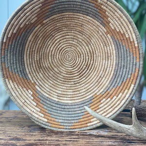 May include: A round, coiled basket featuring a woven pattern in shades of tan, light brown, and muted gray. The basket has a deep, bowl-like shape with concentric circular stitching radiating from the center to the wide, open rim.