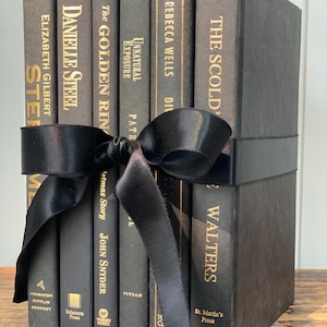 May include: A collection of dark gray books tied with a black satin ribbon. The book titles include "STE", "DANIELLE STEEL", "The GOLDEN RIN", "Unnatural Exposure", "REBECCA WELLS", and "THE SCOLD". The books are stacked on a wooden surface.
