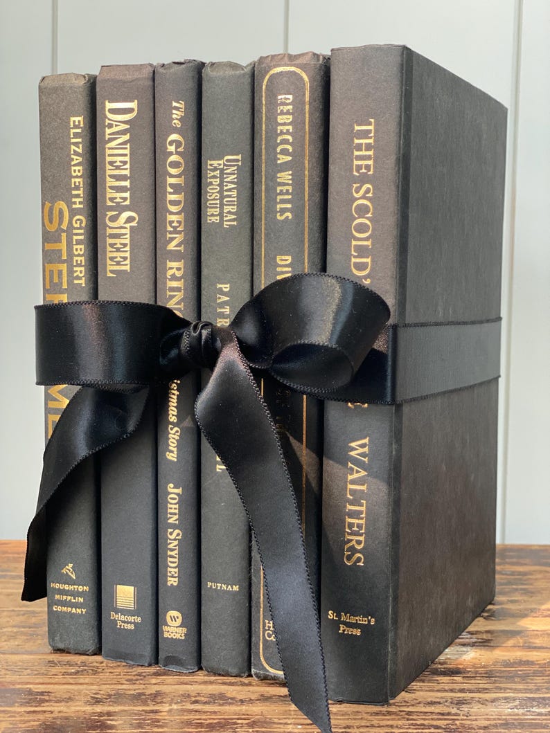 May include: A collection of dark gray books tied with a black satin ribbon. The book titles include "STE", "DANIELLE STEEL", "The GOLDEN RIN", "Unnatural Exposure", "REBECCA WELLS", and "THE SCOLD". The books are stacked on a wooden surface.