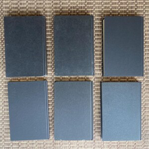 May include: Six hardcover books with dark blue covers are arranged in two rows of three. The books are rectangular and appear to be of similar size. The background is a woven, light brown surface.