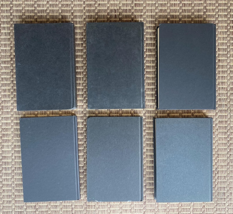 May include: Six hardcover books with dark blue covers are arranged in two rows of three. The books are rectangular and appear to be of similar size. The background is a woven, light brown surface.