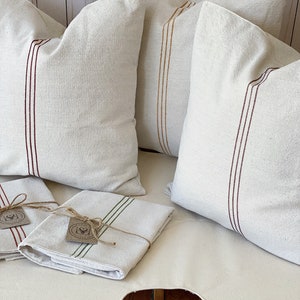 May include: Three off-white square throw pillows featuring vertical stripes in dark red and mustard yellow, accompanied by two folded off-white rectangular textiles with green or red stripes, each tied with twine and a small circular paper tag.