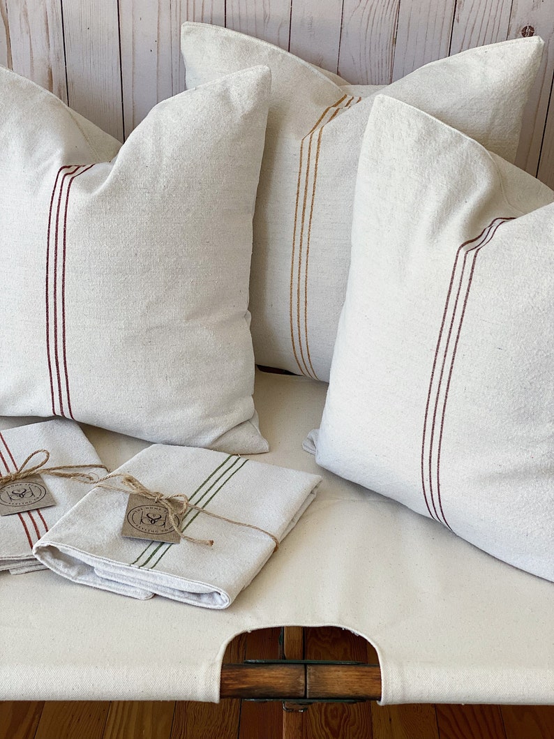 May include: Three off-white square throw pillows featuring vertical stripes in dark red and mustard yellow, accompanied by two folded off-white rectangular textiles with green or red stripes, each tied with twine and a small circular paper tag.