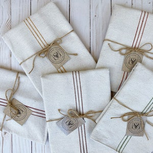 May include: Five folded off-white kitchen towels with colored stripes in mustard, rust, maroon, and green. Each towel is tied with brown twine and features a small, square, tan paper tag printed with the text The Home Spun Ateliers Co.