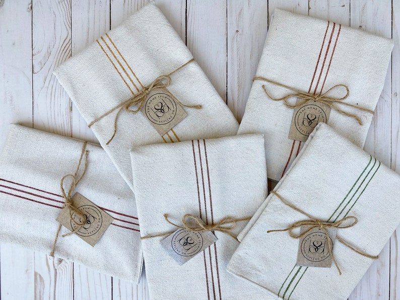 May include: Five folded off-white kitchen towels with colored stripes in mustard, rust, maroon, and green. Each towel is tied with brown twine and features a small, square, tan paper tag printed with the text The Home Spun Ateliers Co.