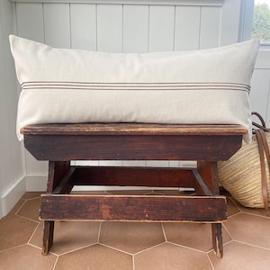 May include: A rectangular lumbar pillow in an off-white fabric featuring two thin, horizontal brown stripes running across the center. The pillow has a smooth, woven texture and is positioned horizontally, showcasing a clean, simple design.