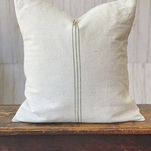 May include: A square throw pillow cover made of off-white, textured fabric. The pillow features a vertical design element consisting of three thin, parallel olive green stripes running down the center of the front panel from top to bottom.