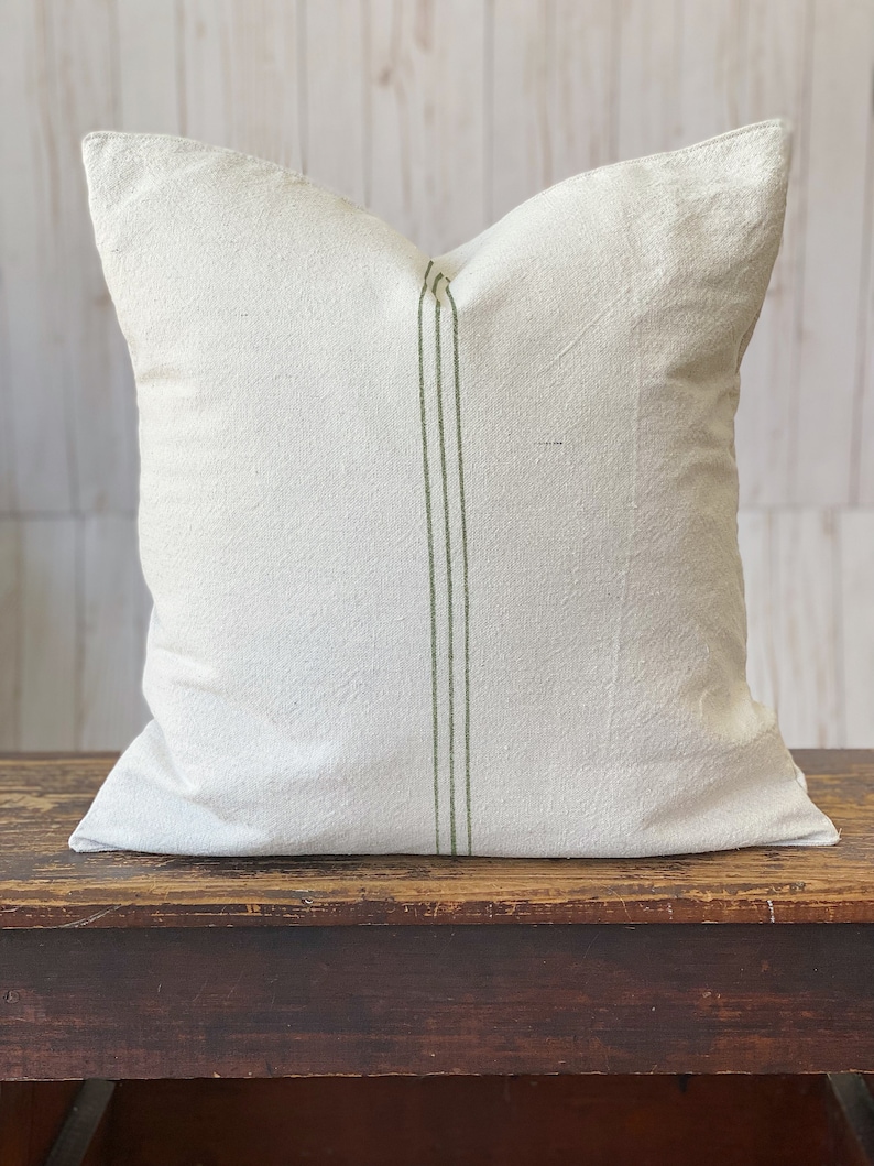 May include: A square throw pillow cover made of off-white, textured fabric. The pillow features a vertical design element consisting of three thin, parallel olive green stripes running down the center of the front panel from top to bottom.
