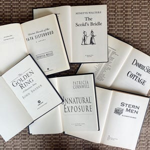 May include: An assortment of open books with white pages and black text, including titles like "Divine Secrets of the Ya-Ya Sisterhood" and "The Scold's Bridle." The books are arranged on a textured, brown surface.