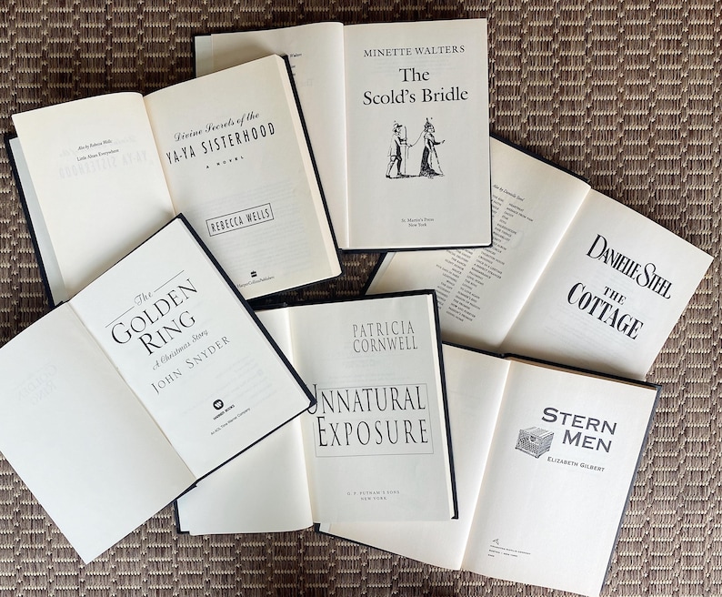 May include: An assortment of open books with white pages and black text, including titles like "Divine Secrets of the Ya-Ya Sisterhood" and "The Scold's Bridle." The books are arranged on a textured, brown surface.