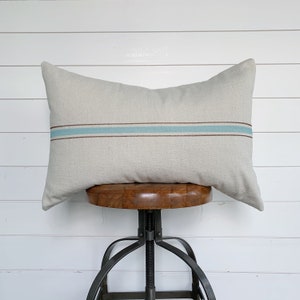 May include: A beige linen pillow with a single blue and brown stripe across the center. The pillow is sitting on a wooden stool with a metal base.