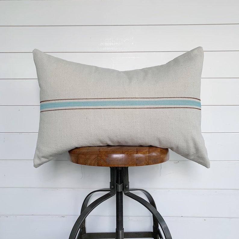 May include: A beige linen pillow with a single blue and brown stripe across the center. The pillow is sitting on a wooden stool with a metal base.